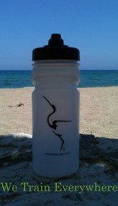 Photo from summer training camp of a water bottle with the logo of the Fujian White Crane Kung Fu & Tai Chi Club on it, standing on a sunny beach in Crete, with the caption "We Train Everywhere"