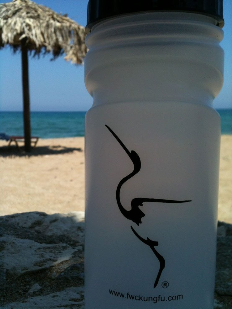 A water bottle with the logo of the Fujian White Crane Kung Fu & Tai Chi Club on it, standing on a sunny beach in Crete, with a beach umbrella in the background.