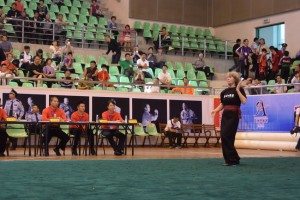 Jill Steen competing in a patterns competition in China in 2015. She is standing on a large green mat, watched by three judges, in a sports auditorium,